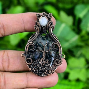 May include: A copper wire wrapped pendant featuring a green and black moss agate stone with a white moonstone accent. The wire is intricately woven to resemble a tree.