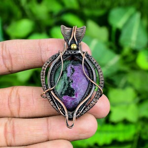 May include: A green and purple gemstone pendant with copper wire wrapping. The pendant has a gold bead at the top.