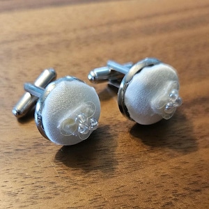 May include: A pair of silver cufflinks with white fabric centers and clear bead accents.