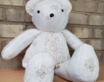May include: A white teddy bear wearing a white lace dress with pearl embellishments. The bear has black eyes and a black nose.