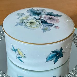 May include: A white ceramic round trinket box with a gold rim and floral design. The lid features a cluster of flowers in shades of yellow, pink, and blue. Additional floral accents are painted on the side of the box.