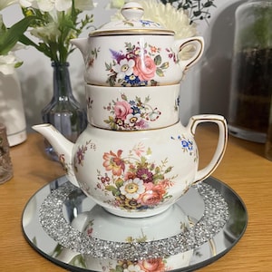 Könnte beinhalten: Ein dreistufiges Porzellan-Teeservice mit floralen Mustern in Rosa, Lila und Gelb. Das Set umfasst eine Teekanne, eine Tasse und einen Deckel, alle mit goldenen Verzierungen. Das Teeservice wird auf einem Spiegel Tablett mit Kristallrand präsentiert.