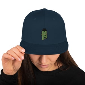 May include: A navy blue baseball cap with a green pixelated face embroidered on the front.