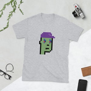 May include: A grey t-shirt featuring a pixelated green character wearing a purple hat. The character has blue eyes and a serious expression.