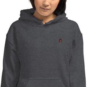 May include: Dark heather gray hoodie with a drawstring hood and a kangaroo pocket. The hoodie features a small embroidered design on the chest. The design appears to be a stylized face with red hair.
