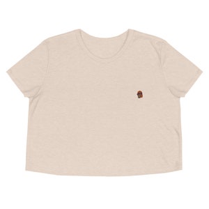 May include: A cropped, light beige t-shirt with a small embroidered image of a person wearing sunglasses.