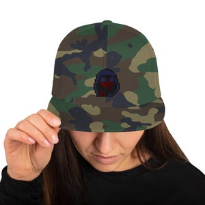 May include: A camouflage patterned baseball cap with a blue and red pixelated character design on the front.