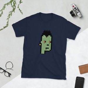 May include: A navy blue short-sleeve t-shirt featuring a pixelated green face with black hair, red eyes, and a yellow earring. The graphic is centered on the chest. The shirt is made of a soft material.