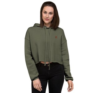 May include: A cropped olive green hoodie with a small embroidered design on the left chest. The hoodie has a drawstring hood and a relaxed fit.