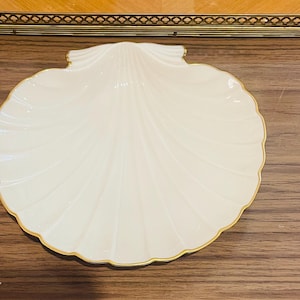 Puede incluir: Plato decorativo en forma de concha, de color crema, con un borde dorado. El plato tiene un diseño acanalado que irradia desde el centro. El plato está sobre una superficie de madera.