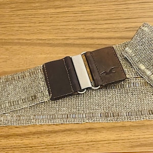May include: A woven belt with a brown leather buckle and a silver-colored clasp. The belt is a neutral color with a textured pattern. The buckle has a logo on it.