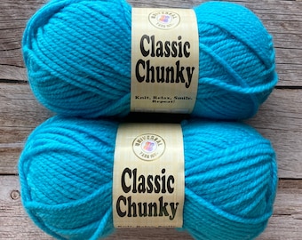 May include: Two balls of turquoise Classic Chunky yarn. Each ball has a label with the text "Classic Chunky" and "Knit, Relax, Smile, Repeat!" The yarn is from Universal Yarn Inc. The yarn is on a wooden surface.