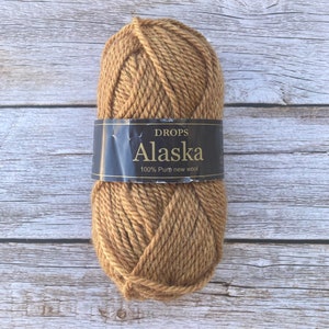 May include: A skein of Drops Alaska yarn in a warm, tan color. The label reads "Drops Alaska 100% Pure new wool". The yarn is tightly wound and appears soft, perfect for knitting or crocheting projects. The background is a light wood surface.