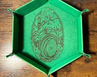 May include: A green, hexagonal dice tray with a detailed illustration of a hobbit hole. The interior is a vibrant green, and the design is etched in a darker shade. The tray has folded corners with metal clasps.
