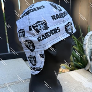 May include: A light gray skull cap featuring a repeating black and white Raiders team logo and the word Raiders printed in bold black text. The cap has visible zigzag stitching along the seams and is displayed on a black mannequin head.