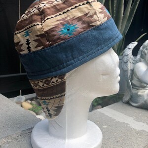 May include: A patchwork cap with a Southwestern-inspired design. The cap features a brown, tan, and turquoise pattern on the crown, with a dark blue denim brim and ear flaps. The cap is displayed on a white mannequin head.