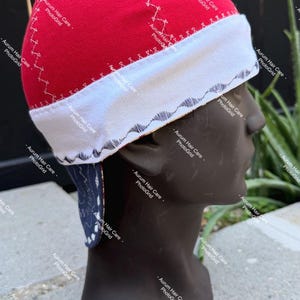 May include: A red skull cap featuring a wide white band with a dark blue wave pattern along the bottom edge. The red section has white cross-stitch detailing, and a dark blue fabric flap with a white paisley print hangs from the back.