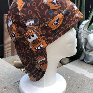 Può includere: Un cappello in tessuto marrone e arancione con un motivo ripetuto di un personaggio di cartoni animati di auto e strumenti. Il cappello è esposto su una testa di manichino bianca. Il cappello ha una tesa curva ed è cucito con un filo a contrasto.