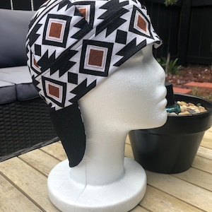 以下が含まれることがあります： 茶色のアクセントが付いた、白黒の幾何学模様の溶接キャップ。キャップには首と肩を覆う長いフラップが付いています。