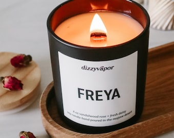 May include: A black candle with a brown glass top, labeled "dizzyvapor FREYA 8 oz sandalwood rose + fresh daisy soy candle hand poured in the mojave desert" is lit and sitting on a wooden coaster.
