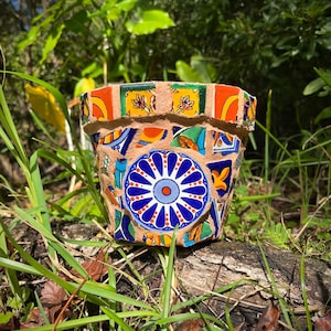 Puede incluir: Una maceta de terracota decorada con azulejos de mosaico de colores. El diseño del mosaico presenta una flor azul y blanca con acentos rojos. La maceta está sentada sobre un tronco en un área cubierta de hierba.