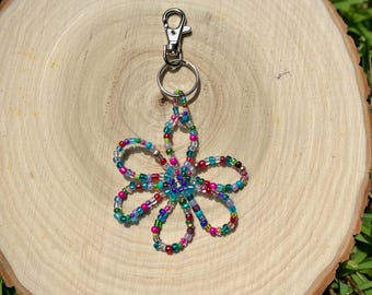 Könnte beinhalten: Ein handgefertigter Schlüsselanhänger mit einer blumenförmigen Perlenverzierung. Er hat einen silberfarbenen Karabiner und Schlüsselring. Die Blume besteht aus bunten Perlen in Rosa, Blau, Grün und Lila. Der Schlüsselanhänger wird auf einer Holzscheibe präsentiert.