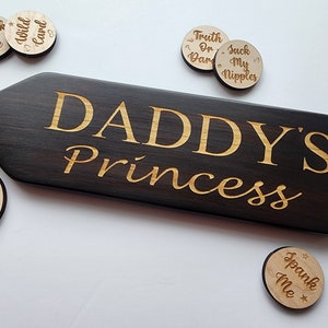 Puede incluir: Una paleta de madera marrón oscuro con el texto "DADDY'S Princess" grabado en letras doradas. La paleta está rodeada de pequeños círculos de madera con varias frases grabadas en ellos, incluyendo "Spank Me", "Wild Card", "Truth or Dare", "Suck My Nipples" y "Midnight Laser".
