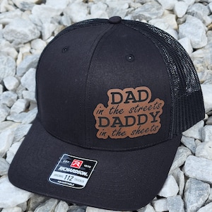 Może przedstawiać: Czarna czapka trucker z brązową skórzaną naszywką z napisem "Dad in the streets Daddy in the sheets". Czapka ma czarny siateczkowy tył i czarny regulowany pasek z logo Richardson.