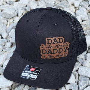 May include: Black trucker hat with a brown leather patch that says "Dad in the streets, Daddy in the sheets". The hat has a black mesh back and a black adjustable strap with a Richardson logo.