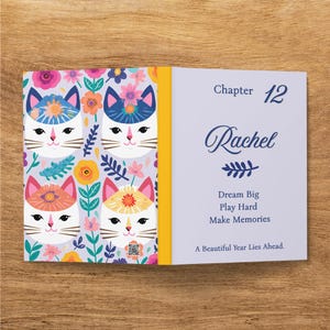 May include: A book with a light purple cover featuring a pattern of white cat faces with colorful floral headpieces. The right side displays the text: Chapter 12, Rachel, Dream Big, Play Hard, Make Memories, A Beautiful Year Lies Ahead.