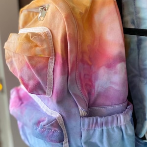 May include: A tie-dye backpack with a white zipper and white trim. The backpack is a light blue, pink, and orange color. The text "BACK TO SCHOOL!" is visible in the image.
