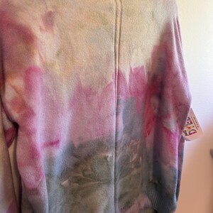 May include: A crew neck sweater with a tie-dye pattern in shades of pink, gray, and green. The sweater has a ribbed collar and cuffs. The dye pattern is most prominent on the lower half of the sweater.