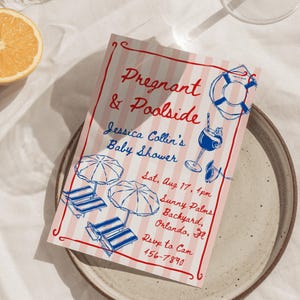 May include: A baby shower invitation with a vintage design. The invitation features pink and white stripes, red borders, and blue illustrations of beach chairs, umbrellas, and a cocktail. The text reads "Pregnant & Poolside" and includes event details.