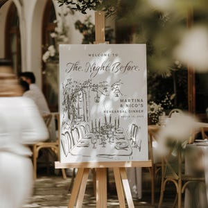 May include: A wooden easel displays a sign with the text "WELCOME TO The Night Before." The sign features a black and white sketch of a dinner table setting. The text "MARTINA & NICO'S REHEARSAL DINNER MARCH 15, 2025" is also visible. The scene is outdoors.