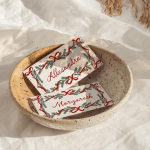 May include: Two rectangular white place cards featuring a festive border of green pine needles and red ribbons. The top card displays the name Allesandra in red script, while the bottom card features the name Margareth in red script, both resting inside a speckled beige ceramic bowl.