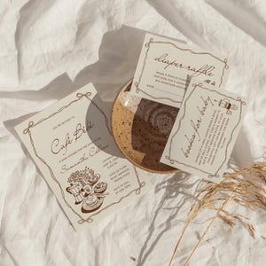 Puede incluir: Invitación a un baby shower con temática de café. La invitación dice "Café Bébé" e incluye detalles del evento. También hay tarjetas para una rifa de pañales y libros para el bebé.