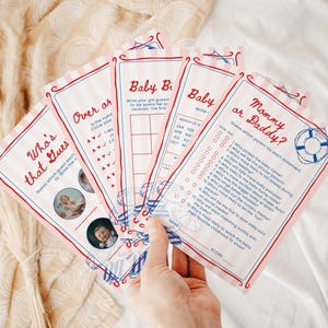 May include: A set of printed baby shower game cards featuring a nautical theme with pink and white stripes, blue life preserver and beach chair illustrations, and red text including titles like Who's That Guess, Over or Under, and Mommy or Daddy.