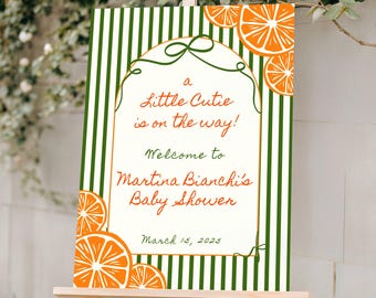 May include: A baby shower sign on a wooden easel. The sign has a green and white striped background with orange citrus fruit illustrations. Text reads "a Little Cutie is on the way!" and "Welcome to Martina Bianchi's Baby Shower" with the date March 15, 2025.