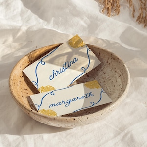 Peut inclure: Deux cartons de placement avec les noms "Christina" et "Margareth" en écriture bleue, ornés d'illustrations de tranches de citron et de lignes bleues. Les cartes sont dans un bol en céramique beige moucheté.