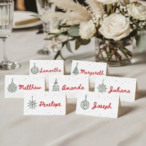 May include: Seven white folded place cards with a speckled pattern, each featuring a green line-art Christmas ornament illustration and a name written in red script. The names include Samantha, Margareth, Matthew, Amanda, Juliana, Penelope, and Joseph.