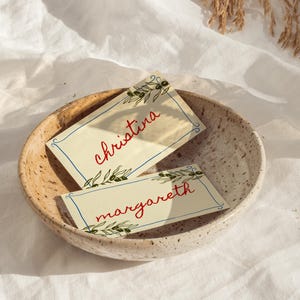 Puede incluir: Dos tarjetas de mesa con los nombres Christina y Margareth en escritura roja, ilustraciones de ramas de olivo y bordes azules, descansan en un cuenco de cerámica moteada. Las tarjetas están sobre un fondo crema.