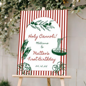Pode incluir: Um cavalete de madeira exibe uma placa com fundo listrado vermelho e branco. A placa diz "Holy Cannoli! Welcome to Matteo's First Birthday" com ilustrações de massa e azeitonas. A data 04.12.25 também está incluída.