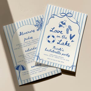 May include: Two rectangular cards with light blue and white vertical stripes. One card features the text Love on the Lake, Nicole's bachelorette party, and illustrations of a bikini, flip-flops, and a life preserver. The other card lists an itinerary.
