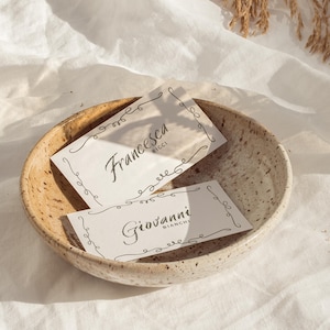 May include: Two white business cards with black script, "Francesca Ricci" and "Giovanni Bianchi", rest in a speckled ceramic bowl. The cards have decorative swirls around the edges. The bowl sits on a white cloth.