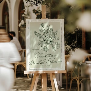 Pode incluir: Uma placa verde clara em um cavalete de madeira com o texto "Welcome to Charlotte's Bridal Shower" e a data de 12 de abril de 2025. A placa apresenta uma ilustração floral preta e uma borda decorativa.