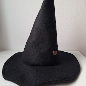 Könnte beinhalten: Ein schwarzer Hexenhut mit breiter Krempe und hoher, spitzer Krone. Der Hut besteht aus einem filzartigen Material und hat ein kleines, rechteckiges braunes Etikett in der Nähe der Basis der Krone. Für Kostüm- oder Dekorationszwecke.