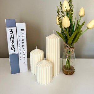 May include: Three white pillar candles with a ribbed design. The candles are arranged on a white surface with a bouquet of yellow tulips in a glass vase.