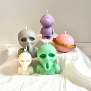 May include: Five colourful candles shaped like aliens and a planet. The candles are made of wax and have a variety of colours, including green, purple, pink, and grey. The candles are all in the shape of aliens with large heads and small bodies. The planet candle is a sphere with a gold ring around it.