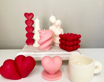 May include: Assortment of Valentine's Day themed candles in various shapes and colors, including a couple holding hands, a heart-shaped mug, and a spiral candle.