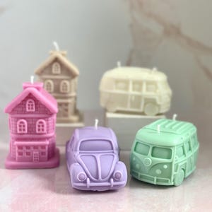 May include: Five decorative candles shaped like houses and cars. The candles are in various colors, including pink, green, beige, and purple.